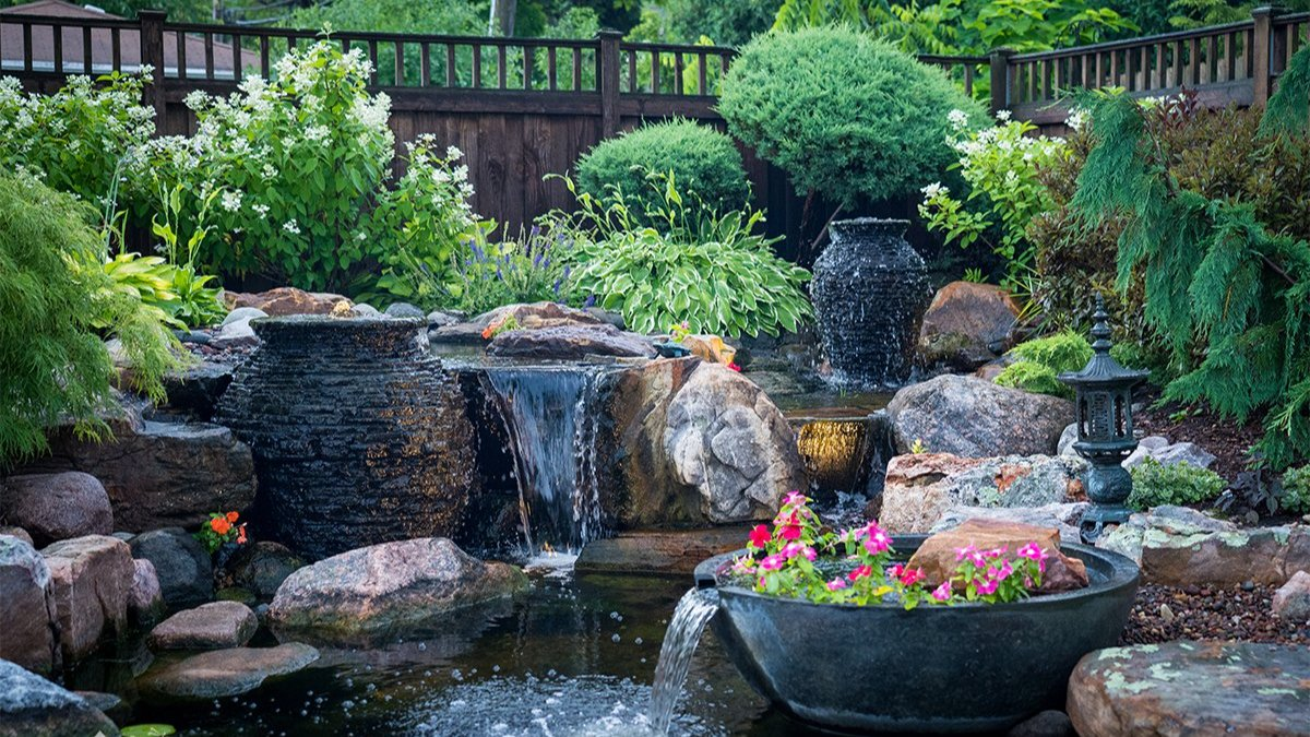 Backyard water feature with cascading waterfall, stacked stone fountains, koi pond, and landscaped garden surrounded by lush greenery and flowering plants